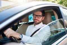 微笑的年轻人戴着眼镜坐在他开车穿过城市的车轮后面