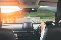 快乐的年轻女子驾驶一辆车与狗沿乡间小路日落在后方视图。女司机手牵方向盘。在旅途中旅行和宠物度假。自由和户外概念