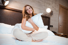 卷曲的微笑妇女坐在一张床上，抱着枕头