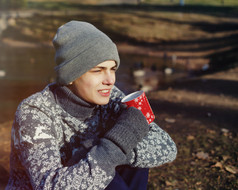 时尚快乐年轻的帅哥穿着毛衣和帽子杯茶或是咖啡在秋天公园附近水