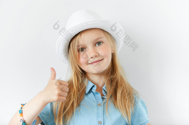 正面可爱的小女婴的肖像，看着相机与自信的微笑，显示竖起大拇指，在白色的墙壁背景上孤立地摆姿势。可爱的学龄前女孩在时髦的衣服