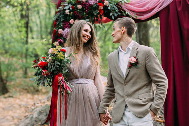 时尚的新娘和新郎手牵着手在婚礼拱门旁边。秋天的婚礼在森林里举行。快乐新婚夫妇