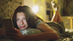 迷人的微笑的女人使用智能手机共享社会媒体躺在床上在晚上在家