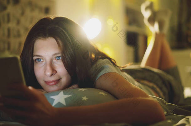 迷人的微笑的女人使用智能手机<strong>共享社会媒体</strong>躺在床上在晚上在家