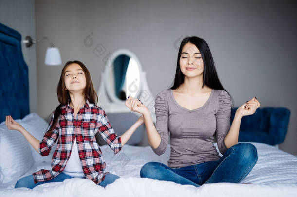 快乐的母亲和女儿打坐和微笑, 而坐在瑜伽姿势在家里的床上.