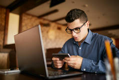 英俊的商人分心的工作在笔记本电脑上看视频的智能手机。自由职业者持有手机和浏览使用高速4g 或5g 互联网。在工作中玩手机游戏的人