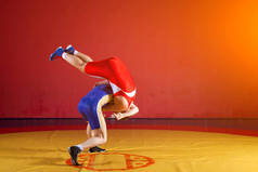 两名身穿红蓝相间制服的男子摔跤选手在体育馆的一块黄色摔跤地毯上摔跤