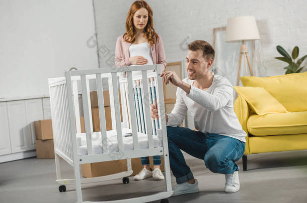 怀孕妇女看着微笑的丈夫固定婴儿床在新公寓