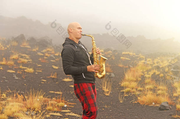 一个秃头男子在一个金色的女低音萨克斯在自然, 反对的背景下, 埃特纳火山火山, 在雾。浪漫的萨克斯爵士乐。这个人是个音乐家。意大利西西里岛.