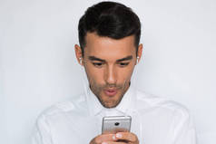 一个英俊迷人的年轻男子在白色衬衫与耳机持有移动电话隔离在浅灰色背景的肖像。播放或搜索应用程序。惊讶的商人 .