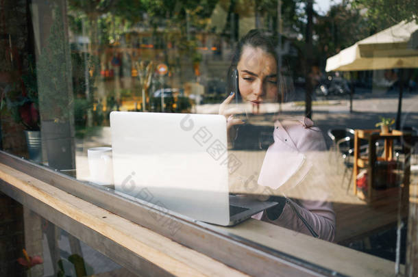 年轻迷人的女性自由职业者呼吁与智能手机, 而坐在舒适的咖啡馆在桌子上的笔记本电脑
