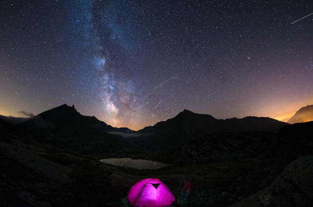 在高空上阿尔卑斯山野营下满天星斗的天空和银河系。在背景前景和雄伟的山峰的照明的帐篷。冒险和探索在夏天.