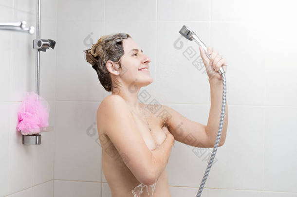 美丽的微笑的女人与洗发水泡沫的头发站在淋浴下