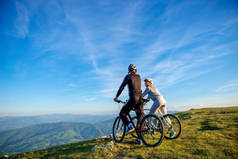 骑自行车的夫妇与山地自行车站在山下的黄昏天空和享受灿烂的太阳在日落.