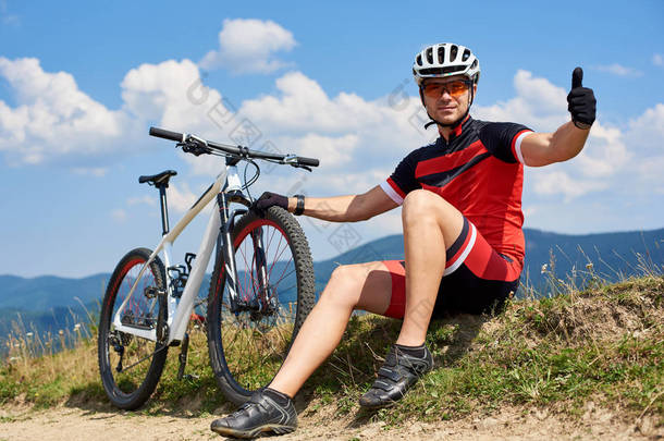 年轻的运动员骑自行车在专业运动服和头盔坐在单车附近的草路边在阳光明媚的一天, 积极的生活方式和户外运动的概念