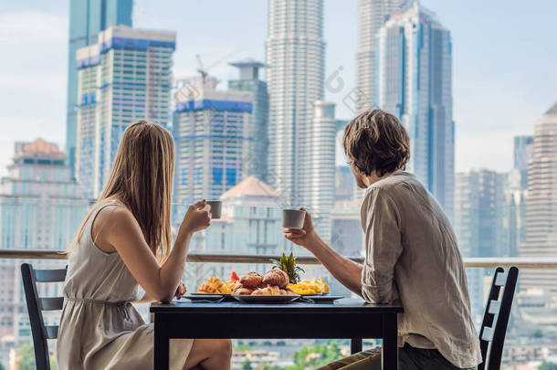 爱的情侣在阳台上吃早饭。早餐桌与咖啡果子和面包 croisant 在阳台反对大都市的背景