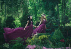 两个女朋友, 一个金发女郎和一个黑发女郎, 手牵着手。背景开花花园。公主们穿着豪华的紫色连衣裙, 长着一列火车, 风吹着一阵风。.