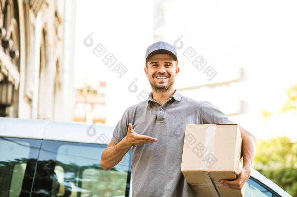 你的船在这里。快乐送货人在灰色衬衫与帽站在街上, 他的纸板箱看着相机与微笑, 在他的送货车前