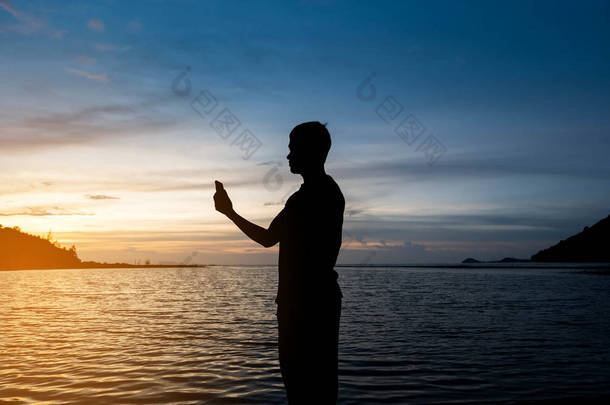一个人独自站在热带海滩上的<strong>剪影</strong>与平静的蓝色海和手持智能<strong>手机</strong>在美丽的黄昏日落。孤独的人概念.