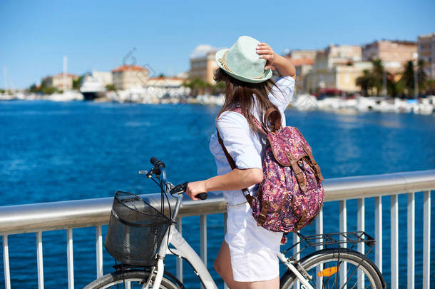 后视图的年轻女子背包和自行车休息附近的围栏石人行道上庇护海港的度假村与巡航摩托艇。旅游、积极的生活方式和度假理念.