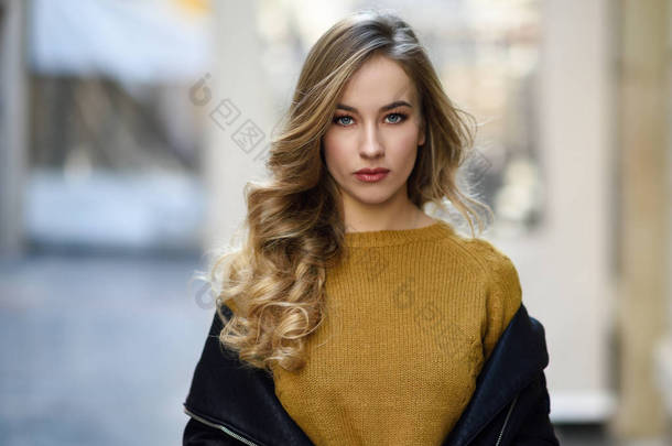 在城市背景下的金发女人。美丽的少女，穿着黑色皮夹克和迷你短裙站在街上。<strong>长长</strong>的卷发发型和蓝眼睛的漂亮的俄罗斯女性.