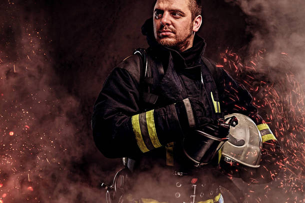 一个穿着制服的职业消防员, 在黑暗的背景下, 在火灾中的火花和烟雾中戴着安全帽.