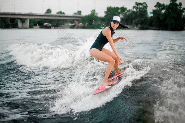 戴着帽子的年轻活跃的黑发女孩在桥和树的背景上骑着摩托艇的 wakesurf