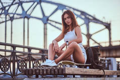 年轻的感官溜冰的女孩穿着短裤和 t恤坐在路堤上美丽的日落.
