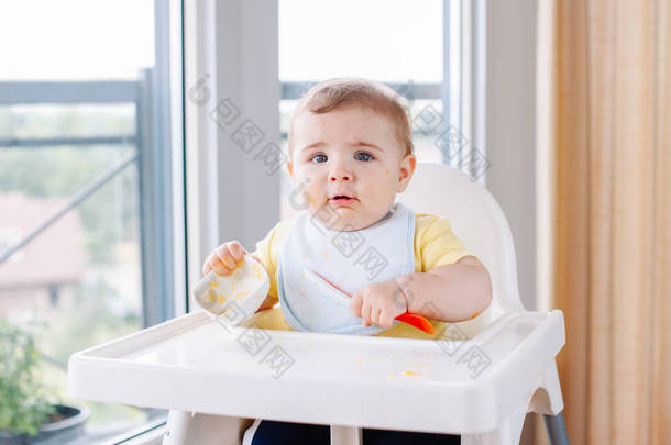可爱可爱的白人孩子的肖像, 肮脏的凌乱的脸坐在高椅吃苹果泥与勺子。每天家庭的童年生活。婴儿尝试补充婴儿食品