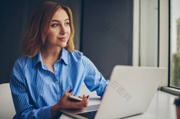在使用无线互联网连接的笔记本电脑上工作时, 她正在用手看着智能手机的年轻女子。沉思的学生对网页设计理念的思考
