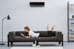 快乐的年轻女子使用智能手机, 而躺在沙发下空调挂在墙上