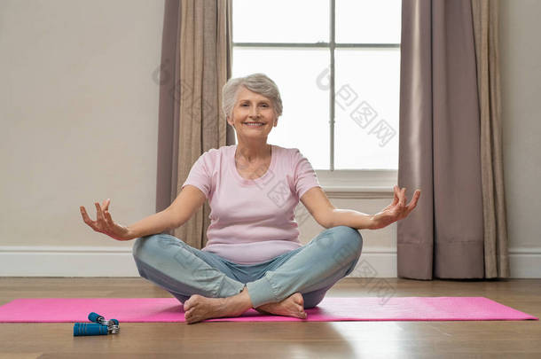 高级微笑的妇女在她的客厅做瑜伽。在家里练习瑜伽的时候, 坐在莲花姿势和冥想的老人放松了。老祖母坐在瑜伽垫上微笑着.