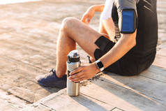 在运动服的亚军男子20s 的裁剪照片与智能手机扶手持有人坐在海边的木码头上, 从热水瓶杯喝水