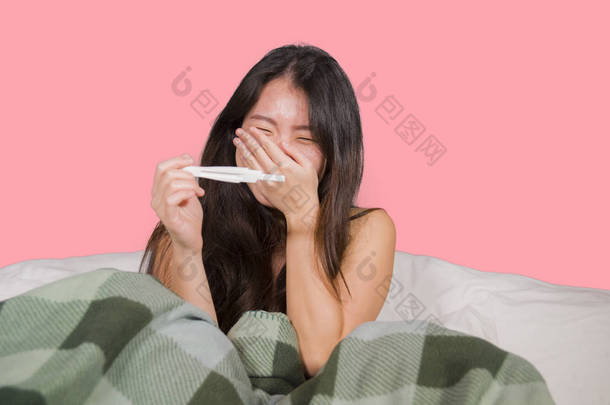 年轻快乐和兴奋的亚洲华人妇女在床上举行妊娠测试检查惊讶阳性怀孕结果微笑欣喜若狂和快乐期待宝宝兴高采烈的感觉祝福