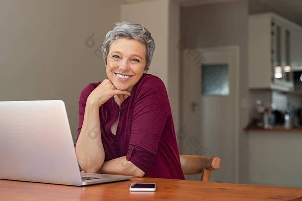 漂亮的高级女士在家里使用笔记本电脑。成熟的微笑的女人看着相机, 而使用电脑。坐在起居室的笔记本电脑的快乐现代退休女士肖像.