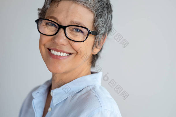 戴眼镜的资深女性肖像, 在灰色背景下看着相机。开朗成熟的商业女性穿着时髦的新眼镜和微笑。成功企业家笑的特写面孔.