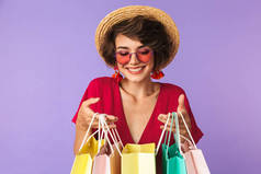 购物狂女人20s 在草帽持有彩色纸购物袋与购买隔离紫色背景的照片