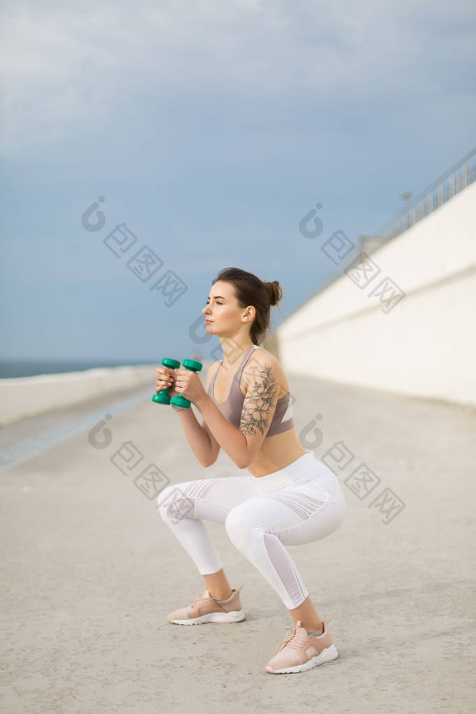 年轻漂亮的女人在运动的顶部和白色的绑腿做蹲在手中的哑铃朦胧地看着一旁的户外