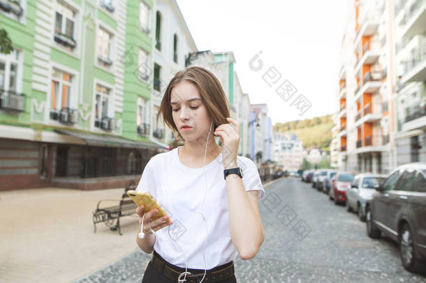 一个严肃时髦的女孩的肖像站在镇上的街道上, 看着她手中的智能手机和听音乐的耳机.