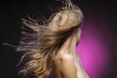 关闭照片的一个转身离开美丽的金发模特女孩裸露的肩膀和飘扬的头发在一个黑暗和粉红色的背景风。干净, 健康的皮肤。广告与商业设计.