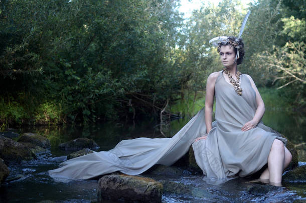 灰色丝绸礼服的美丽的年轻妇女, <strong>羽毛头饰</strong>和海蜗牛项链, 森林仙女, 坐在河石和休息