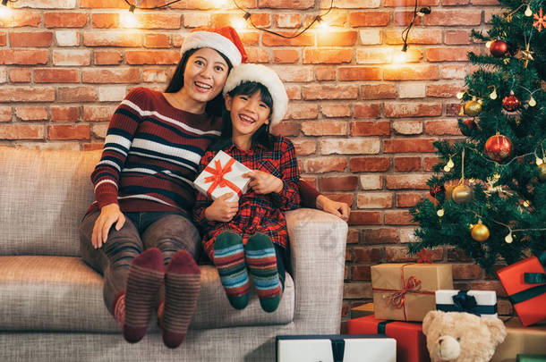 快乐的妈妈<strong>和</strong>孩子坐在沙发上一起庆祝圣诞节一起在客厅里。可爱的小<strong>女孩</strong>拿着礼物显示大露齿微笑。许多礼物<strong>和</strong>玩具周围的<strong>圣诞树</strong>旁边.