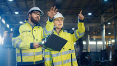 男性和女性工业工程师在通过重工业制造工厂进行讨论和使用笔记本电脑。他们穿着安全帽和安全夹克.