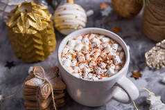 一个白色的杯子, 里面有热可可和棉花糖, 放在白色和金色的圣诞装饰品和冷杉树枝之间的灰色桌子上
