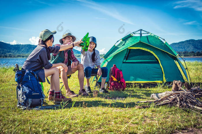 一群年轻的亚洲朋友在湖边享受野餐和聚会, 他们背着野营背包和椅子。年轻人敬酒和欢呼的啤酒瓶。人和生活方式的概念。户外背景主题