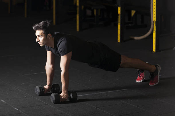 运动员在健身房的哑铃上锻炼俯卧撑。带有复制空间的黑暗摄影概念.