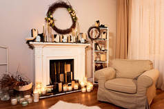 现代设计的宽敞客厅配有秋季装饰。壁炉与蜡烛, 舒适的扶手椅, 秋天的花圈在墙上。背景的夜窗