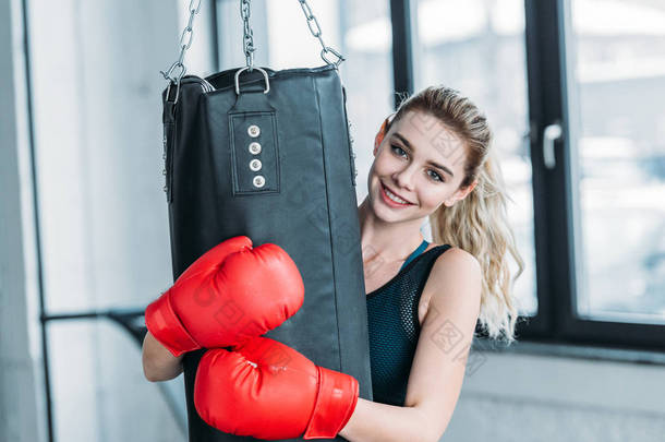 快乐运动的女孩在拳击手套拥抱打气筒和微笑在健身房的相机