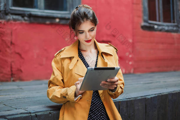 年轻迷人的女人在橙色<strong>风衣</strong>和黑色波尔卡点礼服若有所思地使用平板电脑花时间在老院子里