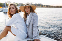 年轻的美丽的女人在白色衬衫和华丽的女士在脱光夹克和草帽梦幻般地看着相机一起在游艇与惊人的海港视图背景